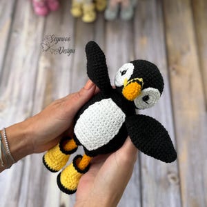 Può includere: Un giocattolo a forma di pinguino lavorato all'uncinetto a mano con corpo nero, pancia bianca e macchie bianche sul viso. Il pinguino ha un becco arancione brillante e piedi arancioni abbinati con bordo nero. Il testo Seynooo Design è visibile in alto a sinistra.