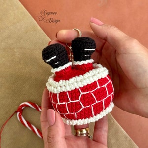 Può includere: Un portachiavi a uncinetto di Babbo Natale con dettagli rossi e bianchi. Babbo Natale è capovolto con le gambe che sporgono da un camino. Il portachiavi è attaccato a un portachiavi argentato.