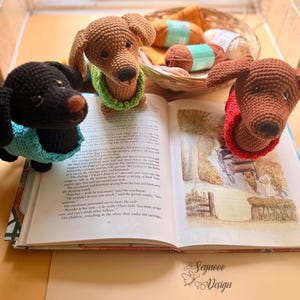 May include: Three crocheted dachshund dog figurines in brown and black, each wearing a different colored sweater. They are positioned on an open book with illustrations, with yarn and a basket in the background. The book has text and illustrations.