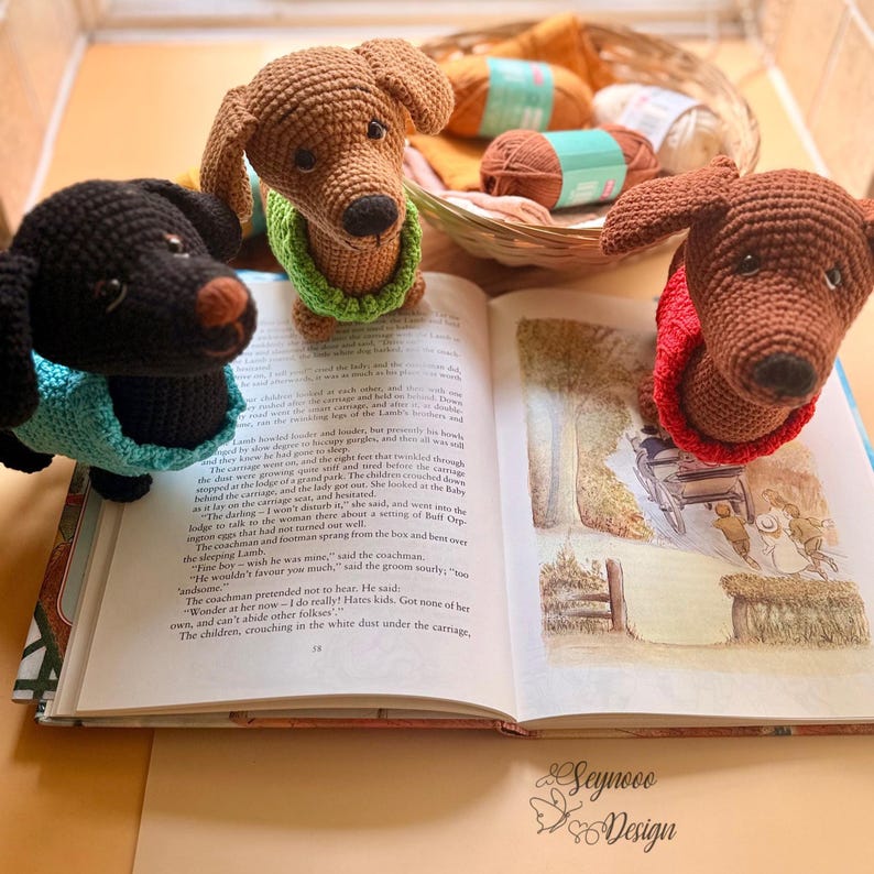 May include: Three crocheted dachshund dog figurines in brown and black, each wearing a different colored sweater. They are positioned on an open book with illustrations, with yarn and a basket in the background. The book has text and illustrations.