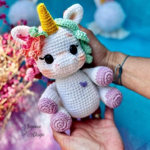 May include: A handmade white crochet unicorn doll with a yellow horn, pink and green mane, and purple heart detail. The unicorn has black eyes and pink cheeks. The doll is being held in a person's hands.