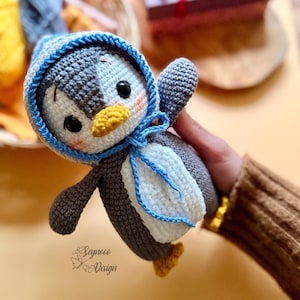 Op de afbeelding: Handgemaakte gehaakte pinguïnpop met een blauw-witte hoed en sjaal. De pinguïn heeft een gele snavel en voeten en zwarte ogen. De pop is grijs, wit en blauw en wordt in een hand gehouden.