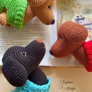 May include: Crocheted dog heads in various colors, including brown, black, and green, are featured in this image. The dogs are positioned near an open book with text. The image also includes the "Seynooo Design" logo.
