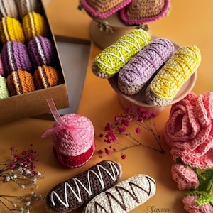 May include: A selection of handmade crochet items, including macarons, eclairs, hearts, and a rose. The macarons are in a box, and the eclairs are decorated with white icing. The hearts are pink and yellow, and the rose is pink and green.
