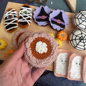 May include: A collection of crocheted Halloween-themed food items, including a pumpkin pie, a ghost, a mummy, and a witch's hat. The items are made with various colors of yarn, including orange, white, black, and purple.