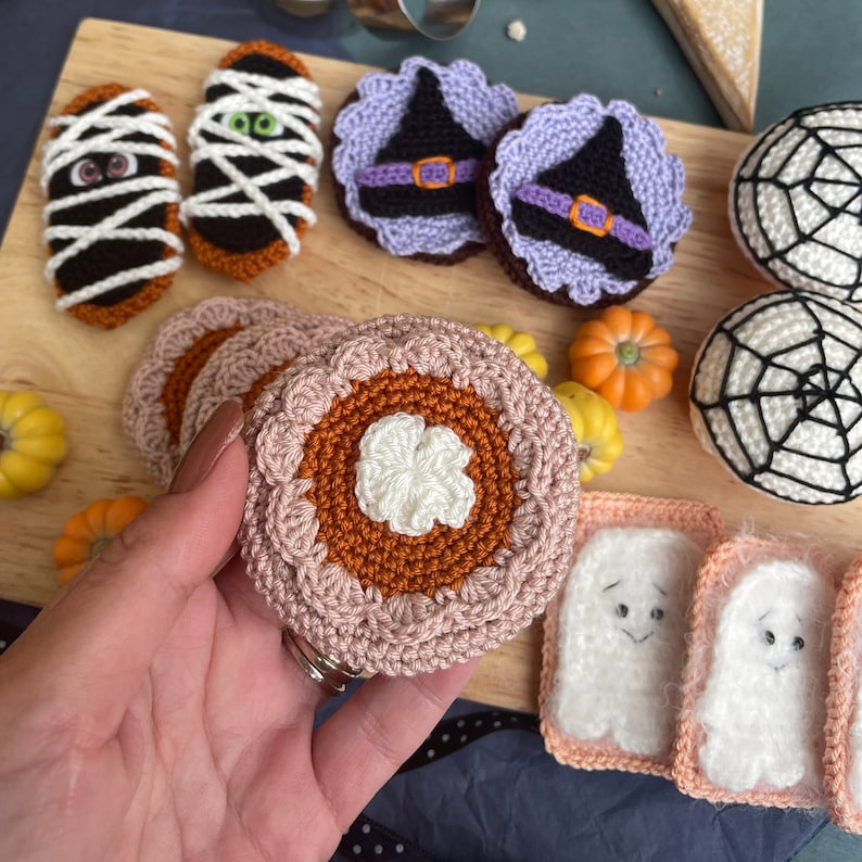 May include: A collection of crocheted Halloween-themed food items, including a pumpkin pie, a ghost, a mummy, and a witch's hat. The items are made with various colors of yarn, including orange, white, black, and purple.