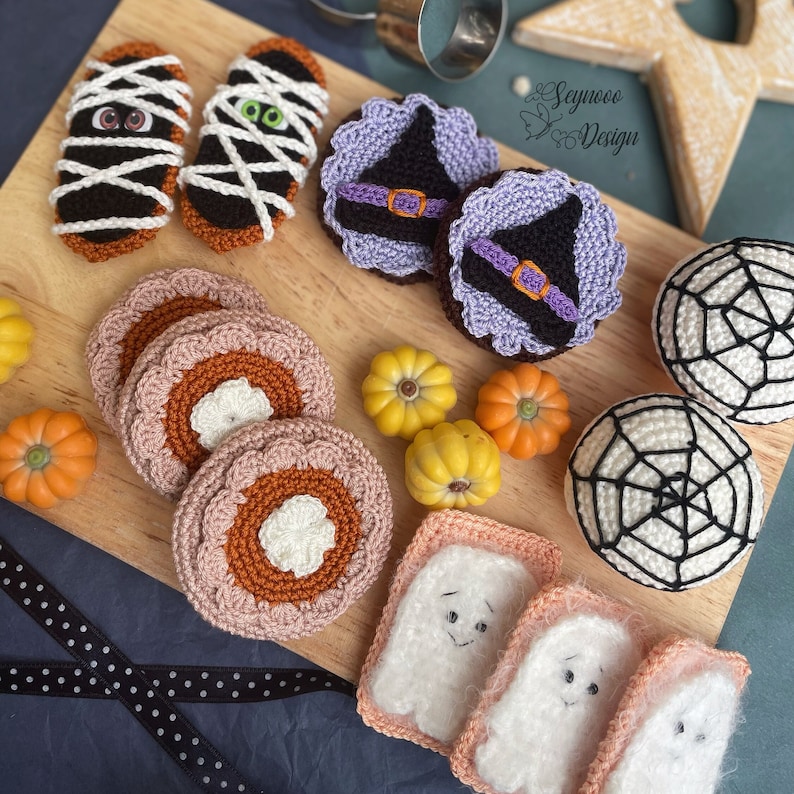 May include: A collection of crocheted Halloween-themed decorations, including ghosts, pumpkins, witches, and spiders. The decorations are displayed on a wooden cutting board with a black and white polka dot ribbon.