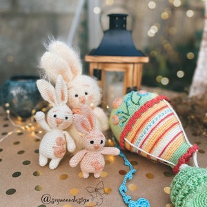 May include: Three crocheted bunny plushies in white, pink, and cream colors, with embroidered details. A crocheted hot air balloon with a green basket is also featured. The plushies and balloon are on a brown surface with gold polka dots.
