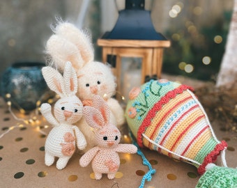 May include: Three crocheted bunny plushies in white, pink, and cream colors, with embroidered details. A crocheted hot air balloon with a green basket is also featured. The plushies and balloon are on a brown surface with gold polka dots.