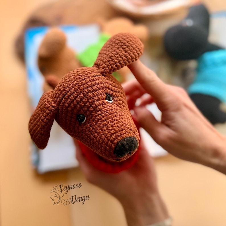 May include: A crocheted brown dachshund dog toy with black eyes and a black nose. The dog is wearing a red collar. The image also shows other crocheted toys in the background.