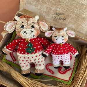 May include: Two crocheted plush toy cows wearing red and white Christmas pajamas. The larger cow has a green Christmas tree on its sweater. The smaller cow is standing on a crocheted mat with candy cane designs.