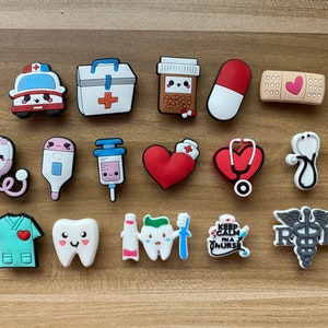 May include: A set of 14 colorful, cartoon-style medical themed shoe charms. The charms include a stethoscope, a heart, a first aid kit, a pill bottle, a bandage, a thermometer, a syringe, a tooth, a pair of scissors, a  ambulance, a  nurse's cap, and a caduceus.