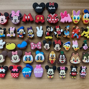 May include: A collection of 30 colorful cartoon character shoe charms featuring Mickey Mouse, Minnie Mouse, Donald Duck, Goofy, and other Disney characters. The charms are made of rubber and have a variety of designs, including classic characters, holiday themes, and everyday objects.