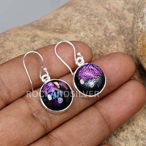 May include: Silver dangle earrings with circular dichroic glass pendants. The pendants display a blend of purple, black, and white hues. The text "ROCKANDSILVER" is visible on the earrings.