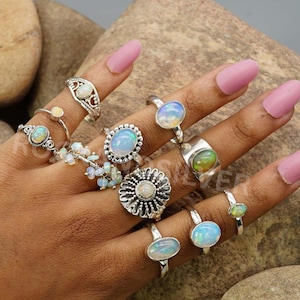 May include: A hand showcasing a selection of opal rings. The rings have diverse designs, including oval, round, and floral settings. The opals exhibit a spectrum of colours, from milky white to iridescent shades. The rings are made of silver.