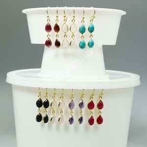 May include: A selection of gemstone earrings with gold-tone hooks. The earrings showcase teardrop-shaped stones in diverse colours, including red, white, turquoise, black, pink, and purple. The earrings are displayed on a white container.