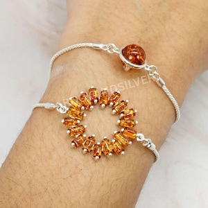 May include: Two silver bracelets with amber-colored gemstones. One bracelet features a teardrop-shaped amber stone, while the other has a circular design of amber stones. The bracelets are on a light background.