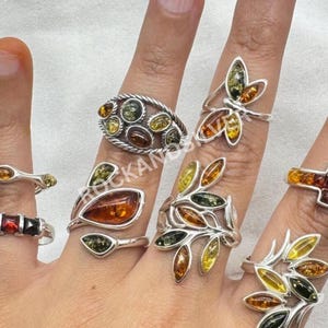 May include: A hand showcases several silver rings, each set with amber gemstones in shades of yellow, orange, and brown. The rings feature diverse designs, including floral and geometric shapes, highlighting the amber's natural hues.