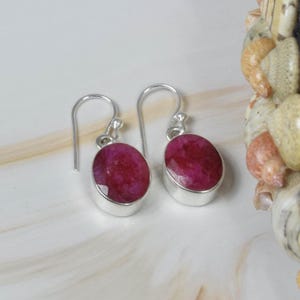 May include: A pair of drop earrings featuring oval-shaped, faceted deep pinkish-red stones set in polished silver bezels. Each earring is suspended from a simple silver hook wire, showcasing a smooth metallic finish and a vibrant, textured gemstone surface.