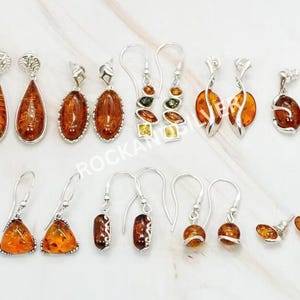 May include: A collection of amber earrings in various designs. The earrings feature silver hooks and settings, with amber stones in teardrop, oval, and geometric shapes. The amber hues range from light to dark orange.