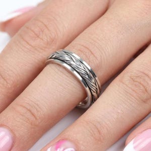 May include: A silver-toned metal ring featuring a central band with a detailed, raised braided texture. The ring has smooth, polished edges on both the top and bottom, creating a distinct contrast with the intricate, woven-style pattern in the center.