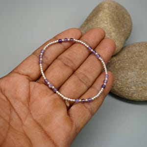 May include: A delicate beaded bracelet featuring alternating small metallic silver spheres and smooth, round purple amethyst gemstones. The circular jewellery piece includes a tiny, circular metallic charm dangling from the bottom of the strand.