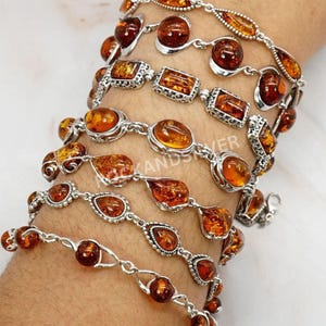 Peut inclure: Plusieurs bracelets en argent ornés de pierres d'ambre. Les pierres d'ambre, de formes variées (rondes, ovales, rectangulaires), sont serties dans des maillons en argent. Les teintes d'ambre varient de l'orange clair à foncé.