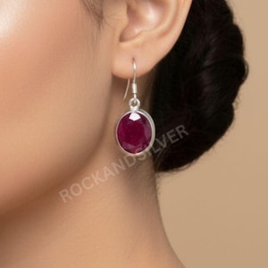 Könnte beinhalten: Ein silberner Ohrring mit einem großen, oval geformten, tiefroten Edelstein. Der Edelstein ist in eine silberne Lünette gefasst und hängt an einem einfachen Haken. Der Ohrring wird am Ohr einer Person gezeigt, mit dem Text "ROCKANDSILVER".