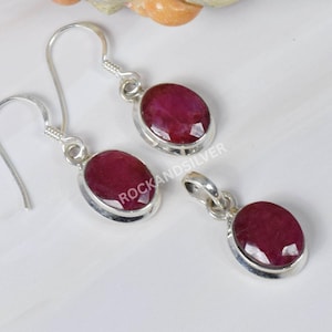 May include: A jewelry set featuring a pair of earrings and a pendant, each with a vibrant red oval gemstone set in a silver metal. The earrings have a hook design, and the pendant has a loop for a chain.