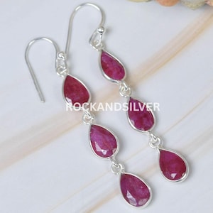 May include: Silver drop earrings featuring three teardrop-shaped, deep red gemstones. The gemstones are set in a silver frame and dangle from a silver hook. The earrings are displayed on a light-colored surface.