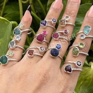 May include: A hand displays several silver rings, each with a unique, raw gemstone. The rings showcase a variety of colours, including green, blue, purple, and red. The gemstones are set in simple, elegant silver bands.