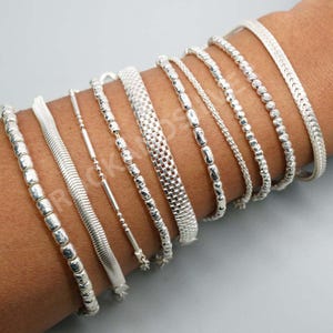Könnte beinhalten: Eine Sammlung von Silberarmbändern in verschiedenen Designs, darunter Perlen-, Ketten- und Webmuster. Die Armbänder werden am Arm einer Person gezeigt, wobei die verschiedenen Texturen und Muster hervorgehoben werden. Der Schmuck ist reflektierend und hell.