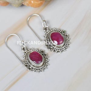 May include: Silver earrings featuring oval, deep pink gemstones. The gemstones are set in an ornate silver setting. The earrings have a hook closure. The text "ROCKANDSILVER" is visible.