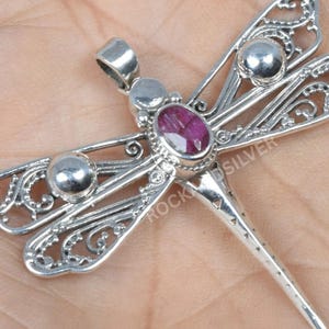 May include: A silver dragonfly pendant with a ruby-colored gemstone at its center. The wings feature intricate filigree detailing and three silver spheres. The dragonfly's tail tapers to a point. The pendant is displayed on a neutral background.