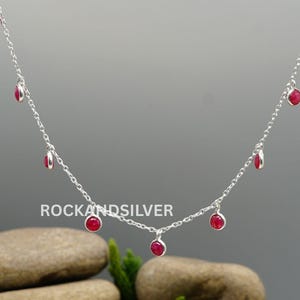 Puede incluir: Un delicado collar de plata con pequeñas piedras preciosas rojas redondas, espaciadas uniformemente a lo largo de la cadena. El collar se muestra sobre un fondo gris neutro, resaltando el contraste entre la plata y las vibrantes piedras rojas. Es una joya.