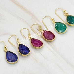 May include: A selection of gold-plated drop earrings featuring teardrop-shaped gemstones in shades of green, red, and blue. The earrings are displayed on a light surface, highlighting the different colours and designs. The gemstones are set in gold-coloured metal.