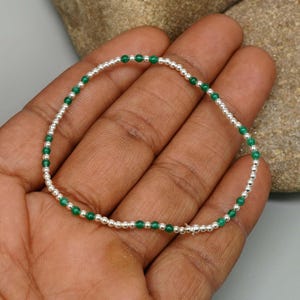 May include: A delicate beaded bracelet featuring a repeating pattern of small, polished silver-toned metallic spheres alternating with vibrant, smooth green circular beads, arranged in a continuous circular loop on a thin, flexible cord.