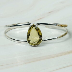 May include: A silver cuff bracelet with a light yellow, pear-cut gemstone. The gemstone is set in a prong setting. The bracelet has a double-band design with a polished finish. The text "ROCKANDSILVER" is visible on the gemstone. The bracelet is approximately 16 cm in circumference.