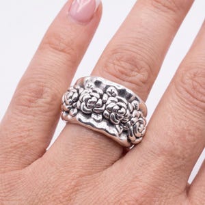 May include: A wide silver ring featuring a continuous band of embossed floral rose motifs. The metal has a polished finish with darkened recessed areas that highlight the intricate details of the sculpted flower petals and small leaves.