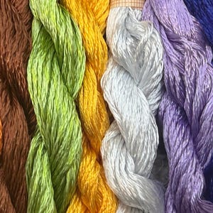 K&ouml;nnte beinhalten: Nahaufnahme von farbenfrohem Stickgarn in Orange, Braun, Gr&uuml;n, Gelb, Wei&szlig;, Lila, Blau und T&uuml;rkis. Die F&auml;den sind eng gewickelt und nebeneinander angeordnet, wodurch eine Vielzahl von Farbt&ouml;nen entsteht.