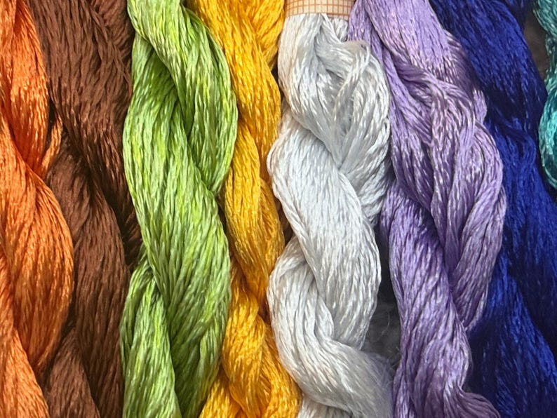 K&ouml;nnte beinhalten: Nahaufnahme von farbenfrohem Stickgarn in Orange, Braun, Gr&uuml;n, Gelb, Wei&szlig;, Lila, Blau und T&uuml;rkis. Die F&auml;den sind eng gewickelt und nebeneinander angeordnet, wodurch eine Vielzahl von Farbt&ouml;nen entsteht.
