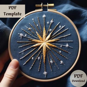 May include: A blue embroidery hoop with a gold and white starburst design. The starburst is made up of gold and white thread and has a small white star in the center. The hoop is on a blue fabric background.
