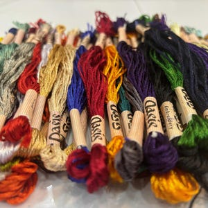Puede incluir: Una colecci&oacute;n de carretes de hilo de bordar en varios colores, incluyendo rojo, azul, verde, amarillo y marr&oacute;n. Los carretes est&aacute;n etiquetados como "Classic Embroidery" y "Daisy".