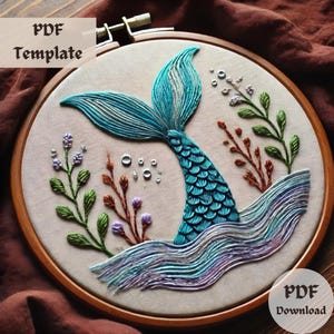 Può includere: Coda di sirena ricamata con filo blu, verde e viola su uno sfondo di tessuto bianco. La coda è circondata da ricami floreali verdi e marroni. Il tessuto è teso su un telaio da ricamo in legno.
