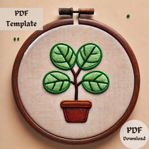 May include: A brown wooden embroidery hoop with a white fabric inside. The fabric has an embroidered design of a green potted plant with five leaves.