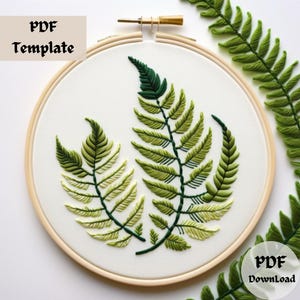May include: Embroidered fern pattern on a white fabric stretched over a wooden embroidery hoop. The hoop is 20 cm in diameter. The fern is green and yellow.