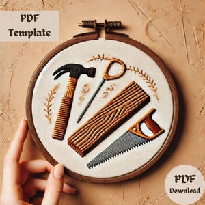 Puede incluir: Diseño bordado de herramientas de carpintería sobre una tela blanca en un aro de bordado de madera. Las herramientas incluyen un martillo, una sierra, clavos y una pieza de madera.