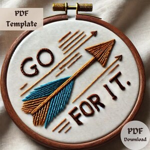 Puede incluir: Arte de bordado en un aro de madera marrón. El diseño presenta una flecha azul y dorada apuntando hacia arriba con el texto "GO FOR IT." en hilo marrón.