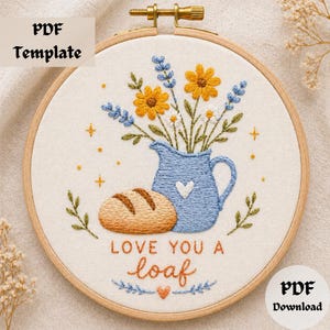 May include: Embroidered hoop art featuring a blue pitcher with a heart, holding yellow and white flowers. A loaf of bread is next to the pitcher. The text "LOVE YOU A loaf" is stitched below. The words "PDF Template" and "PDF Download" are also included.