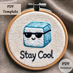 May include: A round embroidery hoop with a white fabric background. The fabric has an embroidered image of a blue cube with a smiling face wearing sunglasses. The text "Stay Cool" is embroidered below the cube.